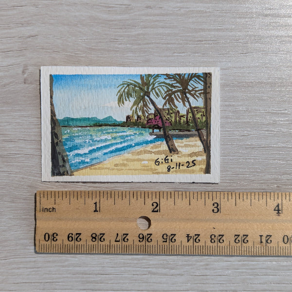 Small beach scene painting with palm trees and ocean, next to a ruler for scale on a wooden surface.